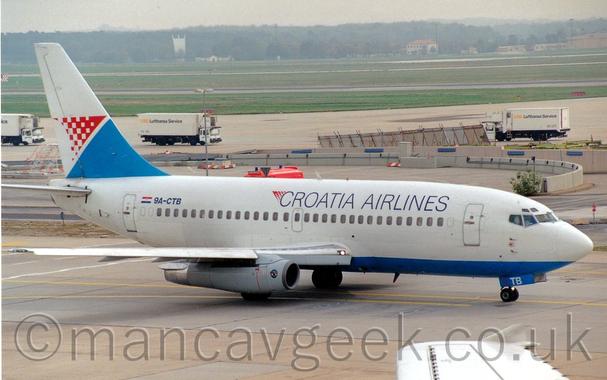 Side view of a twin engined jet airliner taxiing from left to right.
The planer is mostly white with a blue belly, and black "Croatia Airlines"  on the upper forward fuselage.
The black registration 9A-CTB is on the upper rear fuselage, next to a small Croation flag.
Grey concrete apron fills the foreground, with acres of green grass in the background, with trees vanishing in to haze in the distance.
