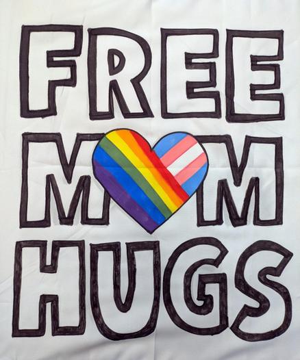 A white bandana with the words "FREE MOM HUGS" outlined in black, and a heart in the place of the "O". The heart is partially in rainbow flag colors and partially in trans pride flag colors.
