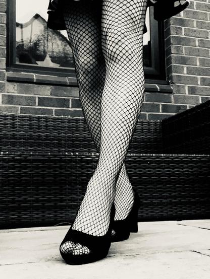 A photo of my legs from about the bottom of my skirt down. My left leg is a little on front of my right making it appear that I’m walking towards the camera. I’m wearing black fishnets tights and black peep-toe heels.