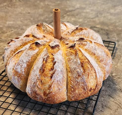 A round loaf of sourdough bread shaped like a pumpkin. Strings are used to tie it into segments and then the dough rises around the strings leaving ridges. There's also some slashes in the dough to create expansion points that add pumpkin details. There's a cinnamon stick shoved in the top which resembles a stem.