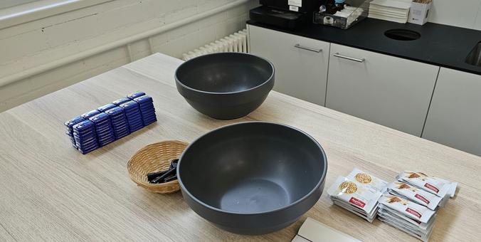 Zwei leere schwarze Schüsseln stehen auf einem hellen Holztisch in einer Büroküche. Daneben liegen ordentlich gestapelt kleine Schokoladentäfelchen in blauen Verpackungen sowie einzeln verpackte Kekse. Eine kleine Korbschale mit schwarzen Faschenöffnern steht ebenfalls auf dem Tisch. Im Hintergrund sind Küchenschränke, eine Kaffeemaschine und ein Abfallloch in der Arbeitsplatte zu sehen.