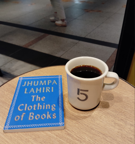 Photo is oa a round light brown wooden table on which the tiny blue paperback book sits. To the right is a off-white coffee mug of black coffee with a 5 on it. In the distance you can see the legs of someone walking passed the cafe inside Tokyo Station