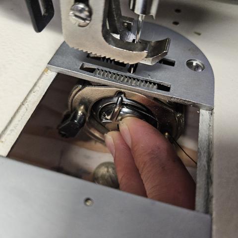a sliding door of a walking foot sewing machine opened to reveal 2 fingers firmly pressing a very fully loaded bobbin into the receiving hole. The aligning pin of the bobbin holder points confidently at the crest of the receiving hole, as there is a knotch, shining glitterously, and poised to receive the aligning indeed.