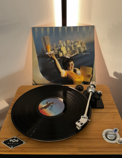 Im Vordergrund dreht sich die Platte von Supertramp auf dem Plattenteller. Dahinter steht die Plattenhülle. Sie zeigt eine lachende Bedienung mit einem Glas Orangensaft vor der Silhouette vom Manhattan, aber mit Frühstücksutensilien.