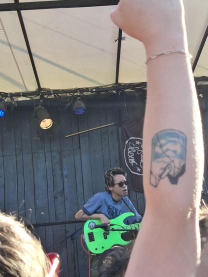 Photo d'un chanteur japonais jouant de la guitare sur une petite scène en extérieur de jour, avec une personne un bras tatoué tendu devant sur la photo.