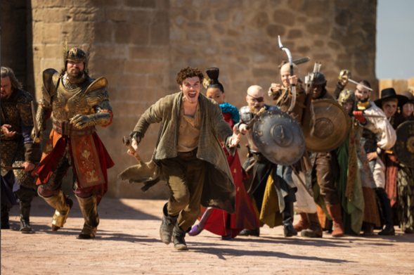 The image depicts a group of people in medieval-style attire running aggressively towards the viewer. At the center, a young man with curly hair is leading the charge, dressed in rugged, brown clothing. To his right, a man in ornate golden armor with a crest is running, carrying a sword. The ensemble includes individuals carrying shields and weapons, each in distinctive costumes that suggest a historical or fantasy setting. The background features a stone wall or fortress, hinting at an ancient or medieval setting under a clear blue sky.