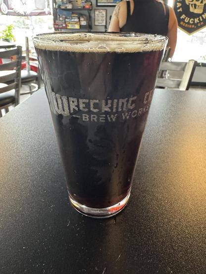 A pint glass filled with a dark beverage sits on a black table, featuring the logo "Wrecking Crew Brew Works." In the background, there is a blurred view of a person and various decorations on the wall.
