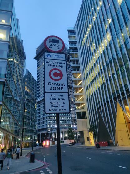 A road sign with a bunch of tall glass office buildings in the background. The sign reads "Transport for London" "Congestion charging Central ZONE" "Mon-Fri 7am-6pm" "Sat Sun & Bank hol Noon-6pm"