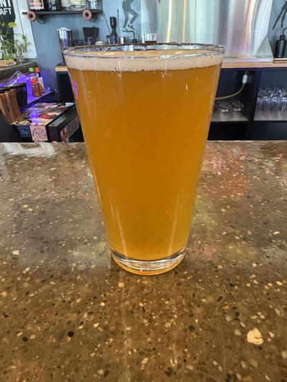 A tall glass of golden-colored beer sits on a textured countertop, with bubbles visible at the top. The background features various bar accessories and bottles.
