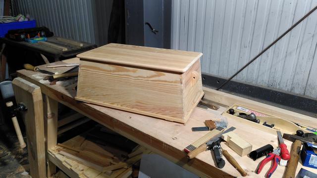 The finished (still unfinished, ie raw wood) chest on the workbench, among various handtools. It is a miniature of a classic sea chest: proud bottom on runners, rectangular planar section, sides as well as front and back slope inwards, wooden chocks for fancywork beckets on the sides, flat panel overhanging lid. The wood is planed and scraped smooth, and has a lovely light blonde color with more creamy brown growth rings. It's reclaimed pitch pine flooring, all slash sawn with flat grain, but so was the original, and that's held up perfectly for close on a hundred years. A few bits of quartersawn larch for flavor.