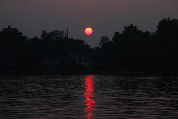 Sunset photo of a red and orange sun in a dark grey sky over a tree-lined lake with the red light of the sun reflecting in the surface of the water.