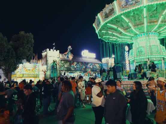 A long shot of the carnival walk with crowds lingering between a haunted dark ride and a swing ride, all lit up at night.