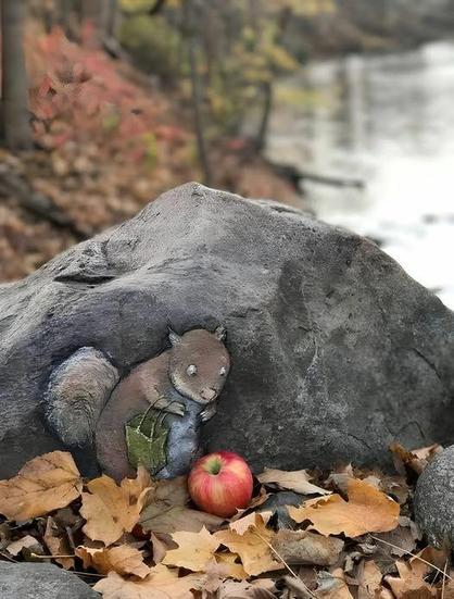 A chalk drawing of a cartoon squirrel on a grey rock. The squirrel has a bag and is bending over to pick up a (real) red apple in the golden autumn leaves