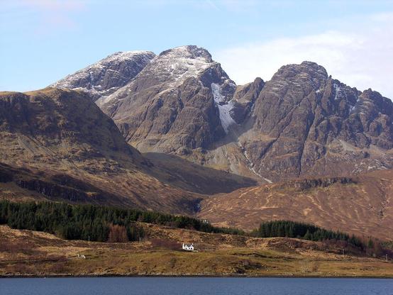 Bla Bheinn. The image shows a view over a loch which is visible across the bottom of the frame. Beyond it the ground rises, with a single white cottage prominent in the distance. Behind that are jagged grey mountains which have their tops covered in a dusting of snow. The scene is in sunlight.