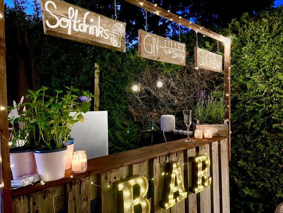 Outdoor bar setup at night featuring a wooden counter with illuminated "BAR" letters at the front. String lights hang above, lighting hand-written signs for "Softdrinks," "GIN - Lillet with berry," and "SEX - Bot." The counter displays several potted plants, candles, an open laptop, and two wine glasses. A lush green hedge forms the background, enhancing the cozy ambiance.