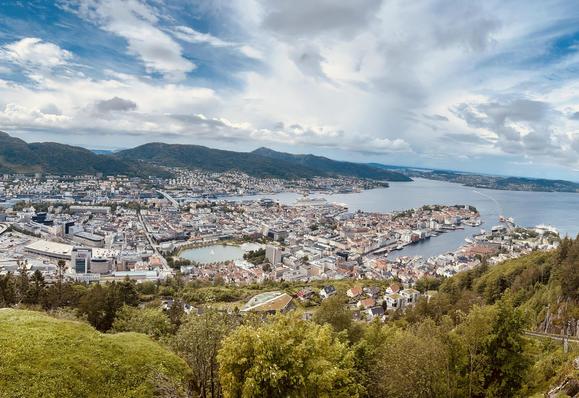 Panoramic view of a coastal city from a hilltop, showing dense urban areas with modern and traditional buildings interspersed by waterways. The city is surrounded by hills and a large body of water, under a partly cloudy sky.