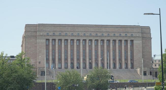 A photograph of a tall, light-colored stone building featuring 14 columns on its front. A wide set of steps lead to them. It is the parliament house of Finland in Helsinki. The picture was taken from afar, so the building is in the background, being car traffic, streets lights, and tree tops.