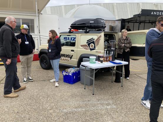Beige Suzuki Jimny coffee truck with black plastic appendages and roof box, rear quarter view. Coffees being served at open-hatch rear.