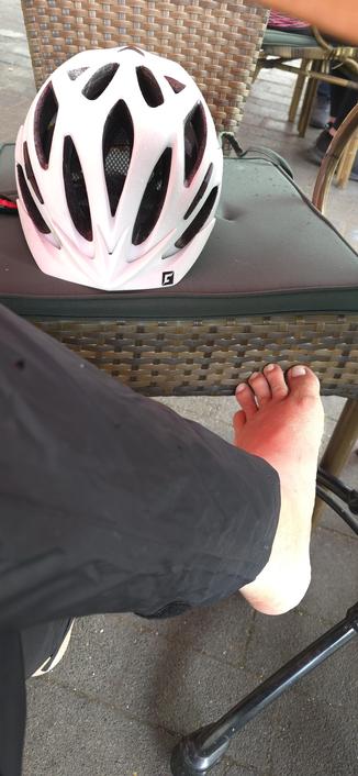 Ein Fahrradhelm liegt auf einen Sessel vor dem ein Unterschenkel in Regenhose und einem nackten Fuß zu sehen ist.
