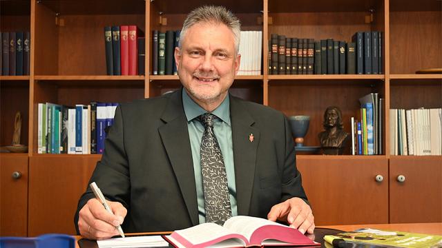 Ein Mann sitzt vor dem Schreibtisch im Bischofsbüro und blickt freundlich in die Kamera. Am Revers seines Jacketts steckt eine Bischofsnadel. Vor ihm liegt ein aufgeschlagenes Buch. In der rechten Hand hält er einen Stift. Hinter ihm ein Bücherregal.