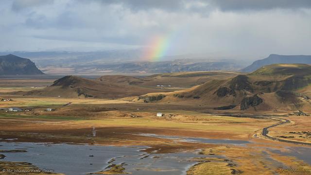 A colour photo of a landscape under a cloudy sky taken from high ground. A black tarmac road, patches of ice, and vehicle tracks can be seen across a foreground of brown grassland. Cloud shadows are dappling the surface. Several small buildings are dotted about, and in the distance is a snow-capped mountain range. In the centre is a small arc of rainbow.