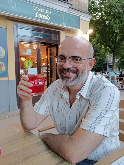 calvo con gafas sonríe, lleva una camisa clara de manga corta, sostiene un vaso de líquido rojo, de fondo un local de hostelería y un árbol a la derecha