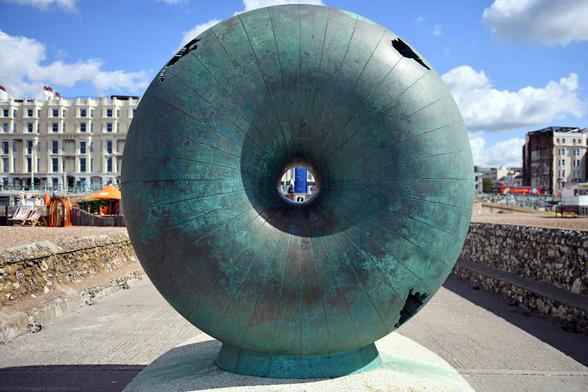 Photo of a bronze, 2.2 tonne torus as a geometric transformation of the world globe and its major continents. Installed 1998 at the end of the Grade 2 listed East Street groyne which was built in 1867 and now popularly renamed as the Doughnut Groyne. City of Brighton & Hove, UK.