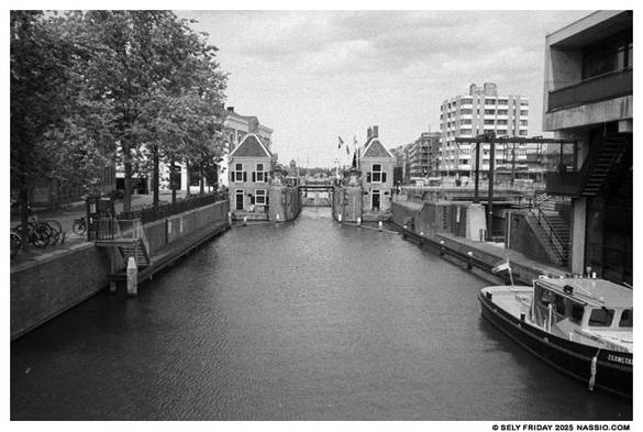 Black and white photograph of the south side of a lock system on the river Zaan. Trees on the left along a road and dock, two buildings on both sides of the entrance to lock and on the right the modern office building.
