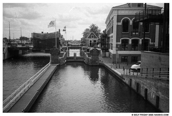 Black and white photograph of the north side of a lock system on the river Zaan. There is a long dock on the left leading to the close lock entrance. To the left of the lock is a small building that houses volunteers of the lock. On the right is a 100 year old building with large windows.