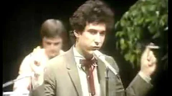 Singer-songwriter Stanard "Stan" Ridgway during a live performance of his band "Wall of Voodoo", playing the cover version of "Ring of Fire" in a television studio. 
He sings into a microphone while he is holding a cigarette in his left hand.