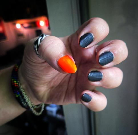 close-up of a hand held up mid-gesture, fingers elegantly curled like they’re about to cast a fabulous spell. Four nails are painted in a deep matte black... bold, velvety, and fierce, catching just enough light to appear almost smoky grey. The star of the show? That unapologetically loud thumb... drenched in pure neon orange, popping like a warning flare thanks to a crisp white base coat beneath. The finish is glossy and wet-looking, almost glowing against the soft skin. A twisted silver ring hugs the thumb, while the wrist flaunts a rainbow bracelet and a silver chain. In the background: a warm, softly lit interior, adding cozy mystery.
