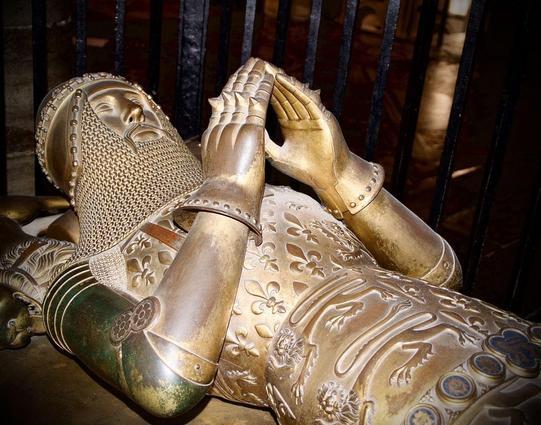Bronze effigy of a medieval knight in full armor, lying supine with hands clasped in prayer. The knight's armor is intricately detailed, featuring floral patterns and chainmail elements. Behind the effigy, metal bars hint at the protective enclosure around the sculpture. The lighting accentuates the metallic texture and ornate design of the armor.