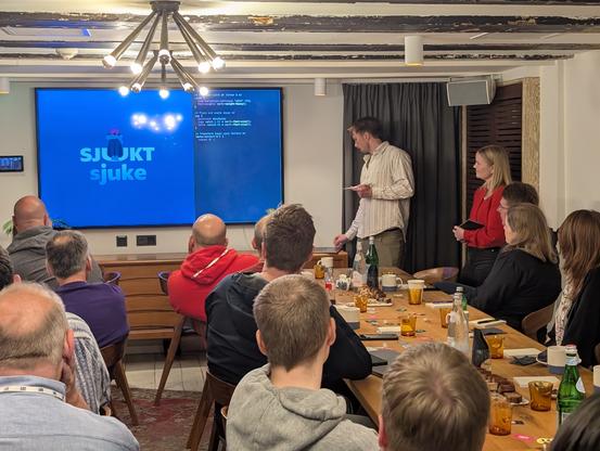 A bunch of people in the café. In the front there are 2 speakers. On the tv there is a visual with text sjukt sjuke and some code next to it.