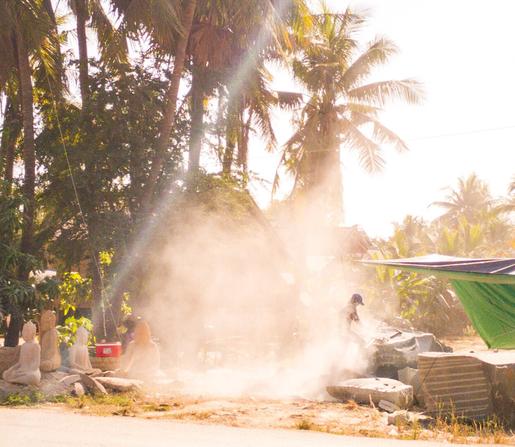 Photo shows a man in a stony dust cloud working on a stone sculpture with a machine tool. It is a sunny day, and the sun plays with the contrasts inside the stone dust. Around the cloud and in the background, there are some sculptures in different modes near finish. To the left there is some large trees, and far in the background there is a low building.  A red fridge box is visible in the shadows under the trees.