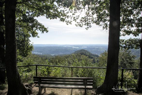 Auf dem Foto sieht man eine Bank, die an einem Aussichtpunkt im Siebengebirge steht, flankiert und beschattet von zwei großen Eichen. Von hier aus schaut mal über eine hügelige Waldlandschaft und den Rhein bis in die Eifel.