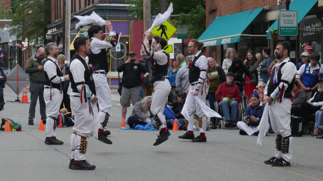 Six morris dancers with white shirts and trousers and black vests, decorated with multicolor ribbons, dancing in a street. Four are standing and two are in motion, feet off the ground and hankies in both hands raised. Behind them is an audience including some other morris dancers.