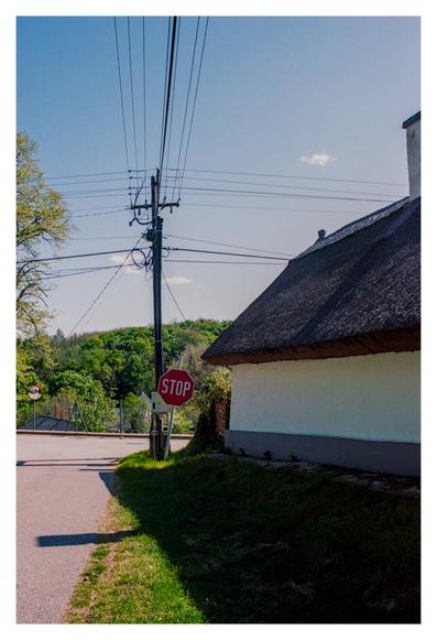 Color Photograph of a rural roadside scene featuring a thatched-roof building with white walls on the right, a red stop sign in the center near a utility pole, and a quiet paved road curving left. The background includes lush green trees and a blue sky with a few wispy clouds.