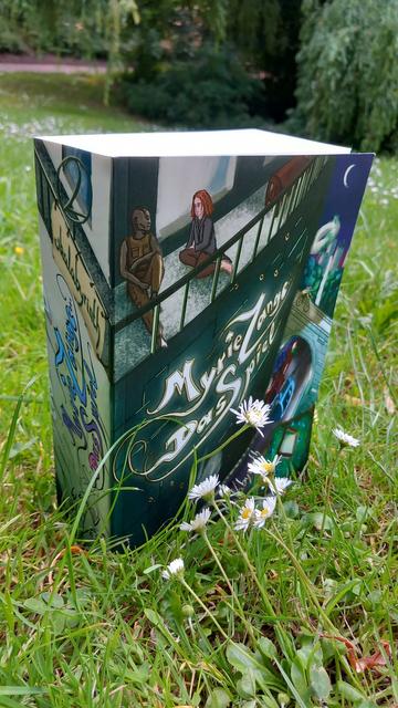 Ein Foto des Buchs "Myrie Zange – Das Spiel" auf einer Wiese im Park. Es steht zwischen Gänseblümchen, im Hintergrund unscharf Weiden. Das Buch ist sehr dick: etwa 6m. Es eigt ein Hochhaus mit Leuchtschrift auf der Fassade, die den Titel buchstabiert. Auf einem Balkon sitzen zwei Figuren. Im Hintergrund ist eine Stadt mit viel Grün und einer Schwebebahn.