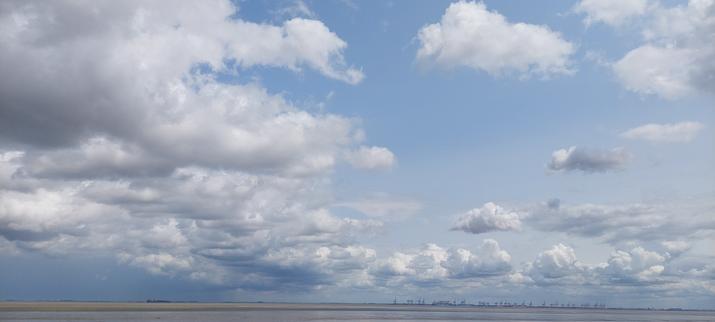 Foto von Küste bei Ebbe bei schönen Wetter, mit paar Wolken, in der Ferne eine andere Küste mit Hafenkrane zu sehen.