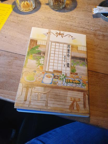 Foto van boek Siwei-straat nr. 4 op een houten cafétafel. Op het omslag het interieur van een Japans huis: houten vloeren, papieren schuifdeur. Op de tafel in het huis allemaal lekker eten.