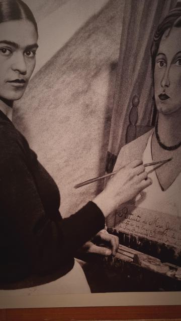 A sepia photograph of Frida Kahlo painting a portrait. She looks at the viewer whilst holding a brush to the canvas.