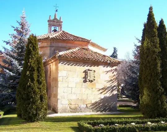 The image appears to depict a small chapel or church, characterized by its stone construction, a tiled roof, and a bell tower. It is surrounded by greenery, including tall trees and possibly gardens, suggesting a peaceful outdoor setting. The architecture features a coat of arms on one side of the building. The clear blue sky implies a bright, sunny day.

Image Credits: Wikimedia / No se ha podido leer automáticamente información sobre el autor; se asume que es Jungpionier (según los derechos de autor reclamados). / Public domain