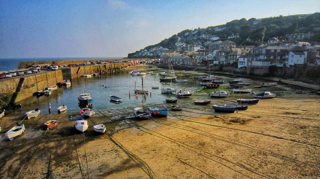 A panoramic view of Mousehole Harbour in Cornwall at low tide. Numerous small fishing boats and pleasure craft are either moored in the remaining water or resting on the exposed sandy harbour floor. To the left, a stone sea wall acts as a car park, with vehicles lining its edge. On the right, a charming village with traditional stone houses climbs up a green hillside under a clear blue sky.