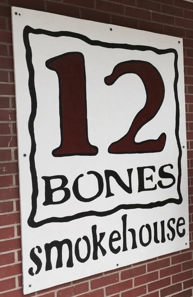 Sign for "12 Bones Smokehouse" on a brick wall. Large red numbers and black text create a rustic feel, evoking a casual barbecue ambiance.