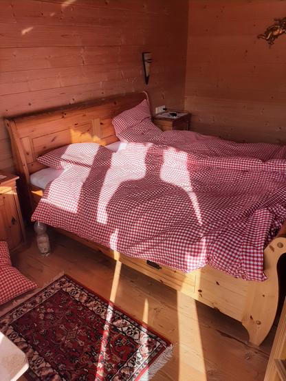 Das Bild zeigt ein gemütliches, rustikales Schlafzimmer mit Holzwänden und einem Holzbett. Das Bett ist mit rot-weiß karierter Bettwäsche bezogen. Die Sonne scheint durch ein Fenster und wirft Schatten auf das Bett und den Boden. Neben dem Bett steht ein kleiner Holztisch mit einem Lampenschirm und ein paar Gegenstände darauf. Die Wände sind mit hellem Holz verkleidet.

Text mit Unterstützung von @altbot