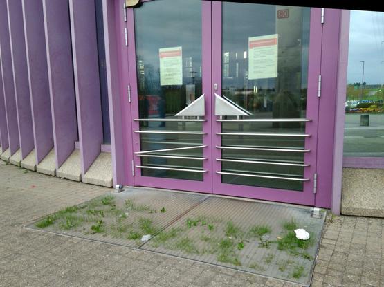 Lila Gläserne Wartehalle des Bahnhof Vaihingen Enz mit Zettel an den Türen, dass gesperrt ist (seit Januar 2022). Vor den Türen wächst etwa 6cm Gras hervor.