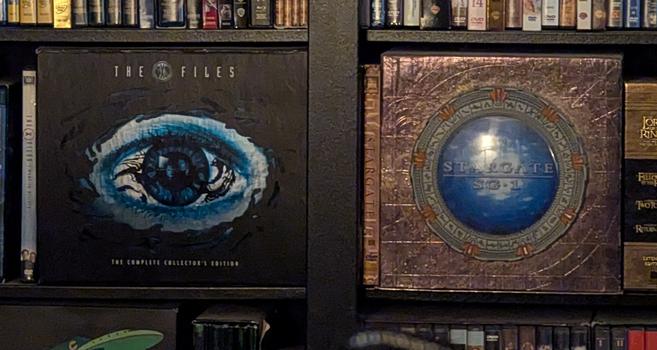 DVD box sets of The X-Files (featuring an eyeball with lids and lashes, and black cancer), next to Stargate SG-1 (featuring the Milky Way Stargate with an active wormhole). The two boxes look like a weird, creepy face