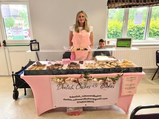 Deelish Cakes stand, a tressle table laid out with about a dozen types of cakes. behind the table is a blonde haired lady with a pink pinefore and a small boy helping out and trying not to eat all the merchandise.