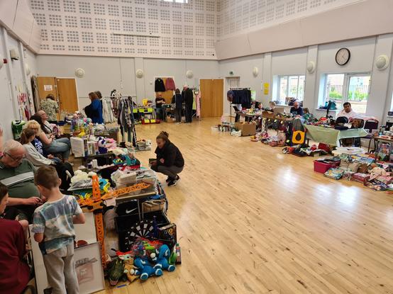 a wide picture of a community centre hall with tables around the edges filled with Brick-a-Brack