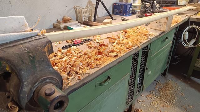 In einem Schraubstock ist ein langes Stück Holz eingespannt.
Überall auf Tisch und Fußboden, liegen Holzspäne und Schnipsel, vom bearbeiten